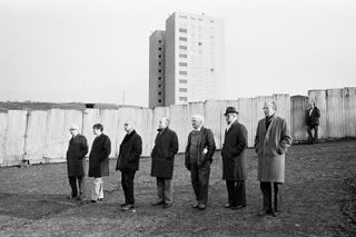 7 older men in coats, stood in a line in front of a fence and a tower block