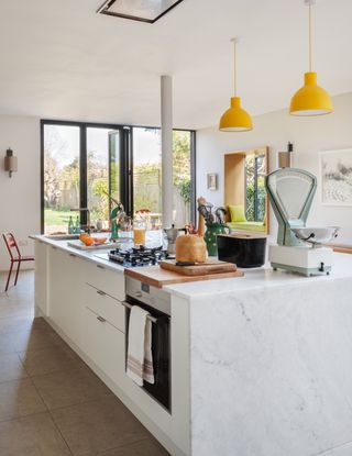 A white kitchen in an open plan living space with a large kitchen island and yellow island lighting