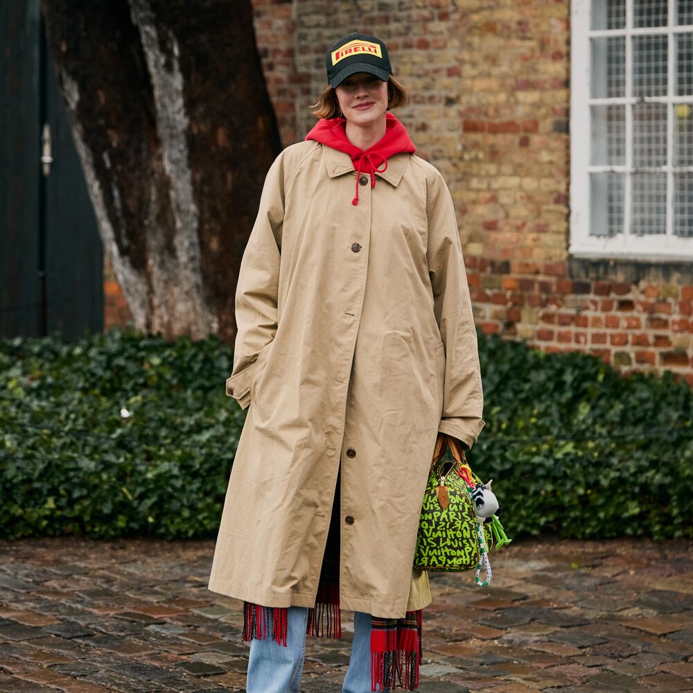A woman wears a trench coat with a red fringe scarf layered under it and a red scarf tied around her shoulders. She also has a trucker hat, bright green Louis Vuitton bag, and cowboy boots. 