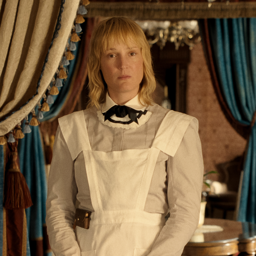 vicky krieps as bridget sullivan wearing a maid's uniofrm standing in a parlor of a victorian era home in a still from monster the lizzie borden story