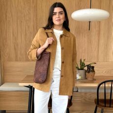 British fashion influencer Anna Howard poses in her modern London kitchen with wood paneled walls wearing a tan suede jacket, burned-out sheer t-shirt, brown woven tote, and white jeans