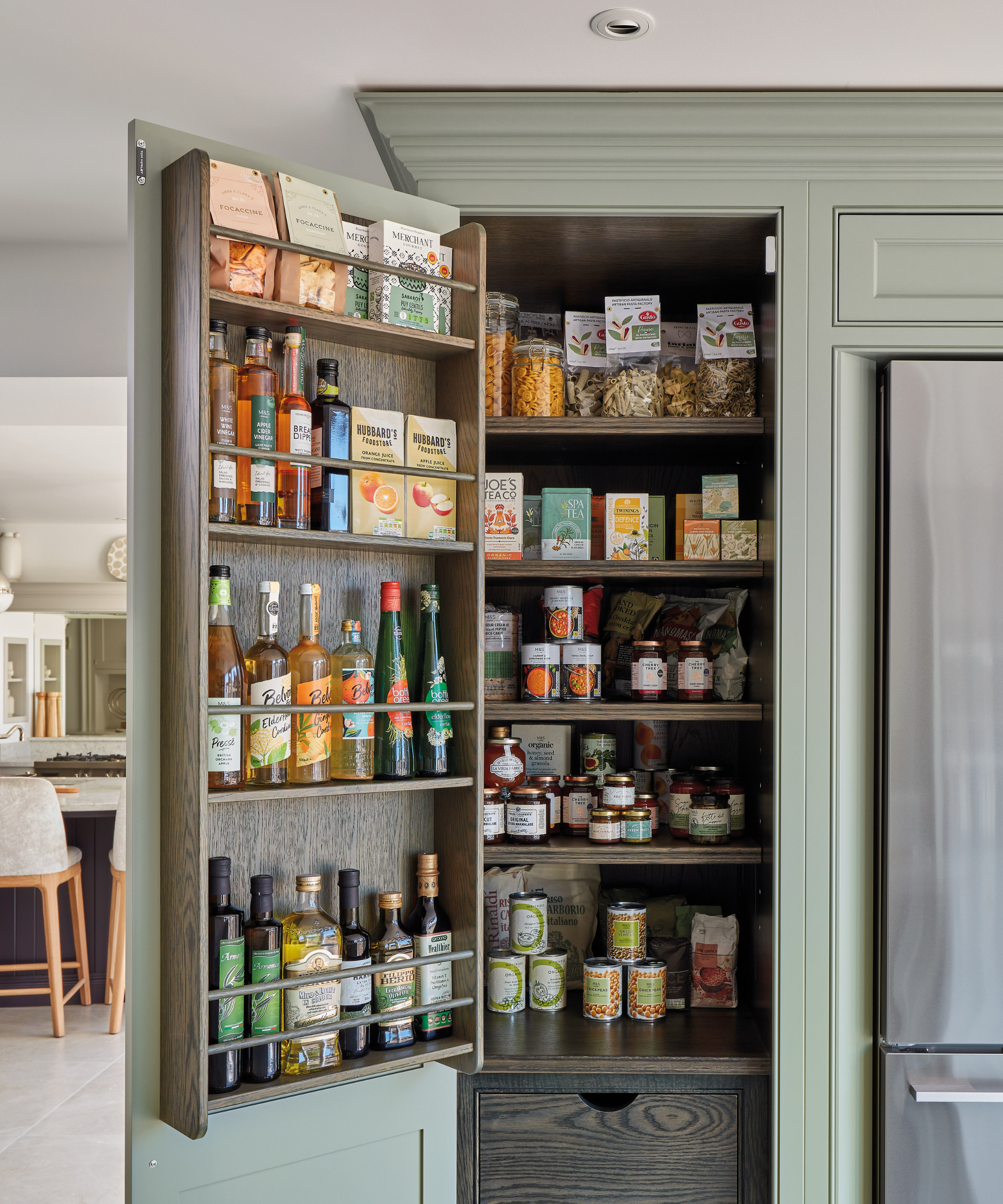 Tall built-in kitchen pantry with sage-green cabinetry, dark wood interiors, and door-mounted shelves, neatly stocked with jars, bottles, pasta, and dry goods for smart, organised storage.