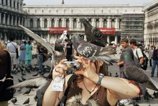 someone taking a photograph with a point and shoot camera in Venice with pigeons all over their head and arms