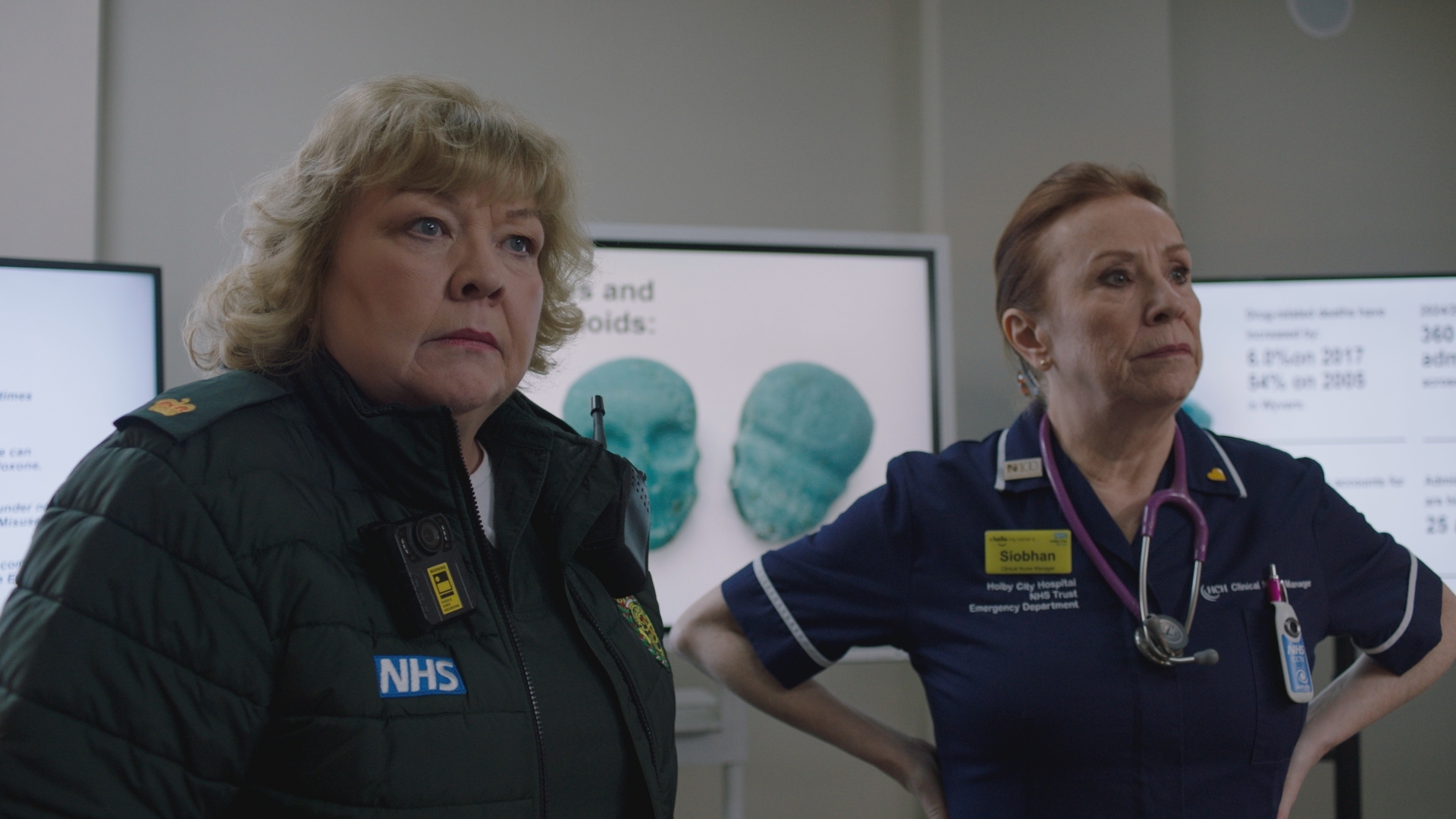 Jan and Siobhan call a meeting with Flynn (out of shot) to persuade him to declare a critical incident. Behind a worried looking Jan and Siobhan are screens with an image of skulls and statistics on them.