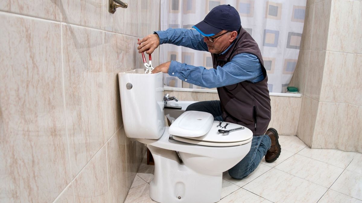 A man with a blue shirt and baseball cap repairing a toilet