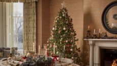 A living room with a tall Christmas tree dressed in ribbons and multi-colored baubles by a mantel with lit tapered candles and a burning fireplace beside a table set with presents, crackers, tapers, plates, and wine glasses