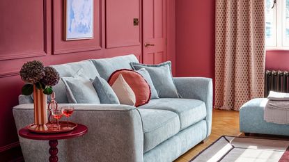 A red painted living room with a light blue sofa, with red cushions on it. A small red side table beside the couch with a tall vase of flowers on it.
