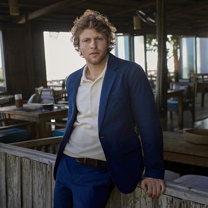 A man wearing Rodd & Gunn blue blazer, jeans and white polo shirt stood next to bar.