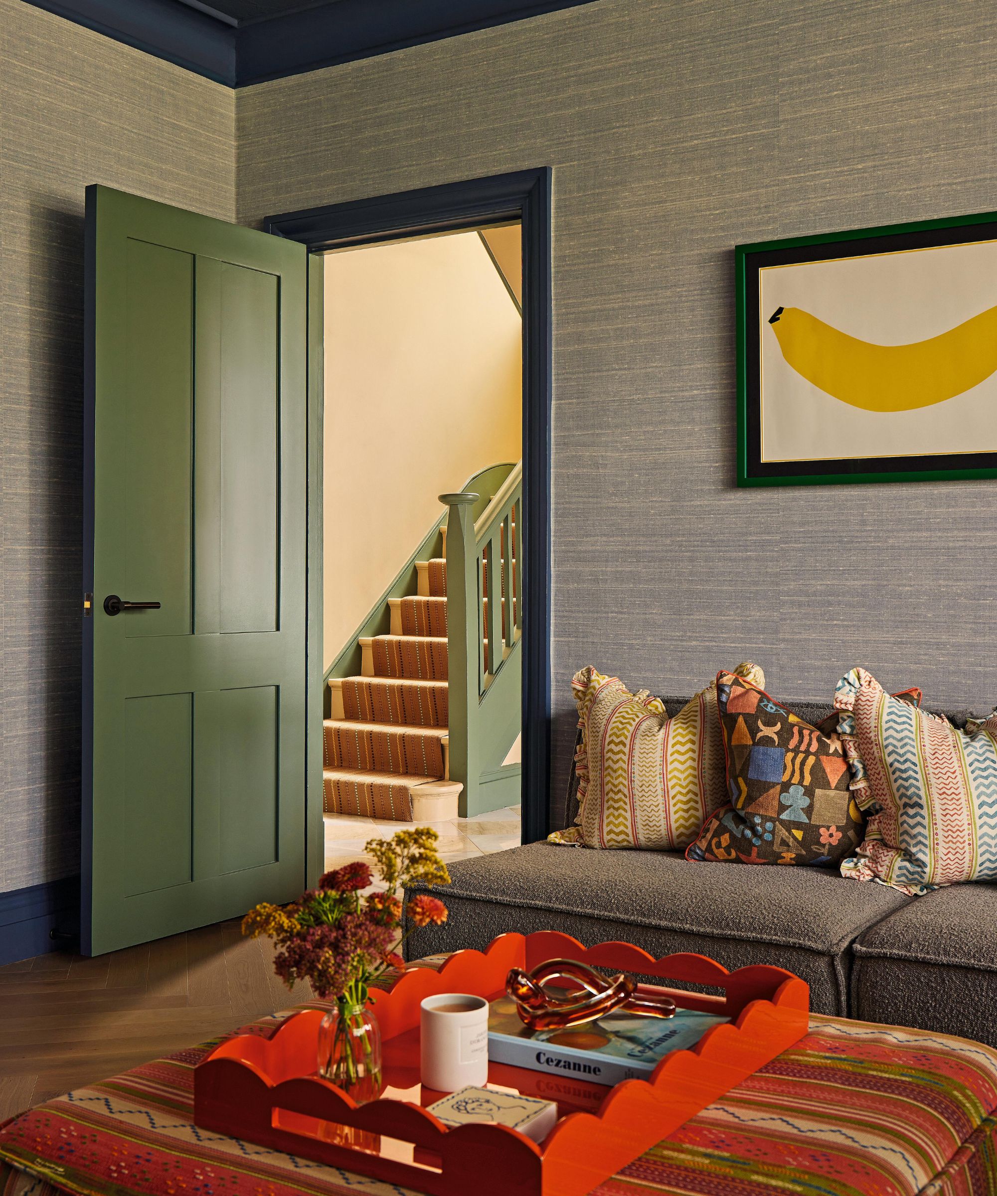 a family room with textured wallcovering, navy painted trim, a green door, grey sofa and orange upholstered ottoman. On the wall hangs a banana painting