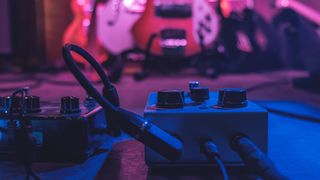 Two guitar pedals in focus with some blurry guitars in the background