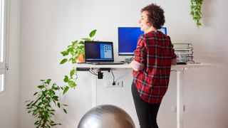 A woman standing and working at a standing desk with a laptop, monitor and plants
