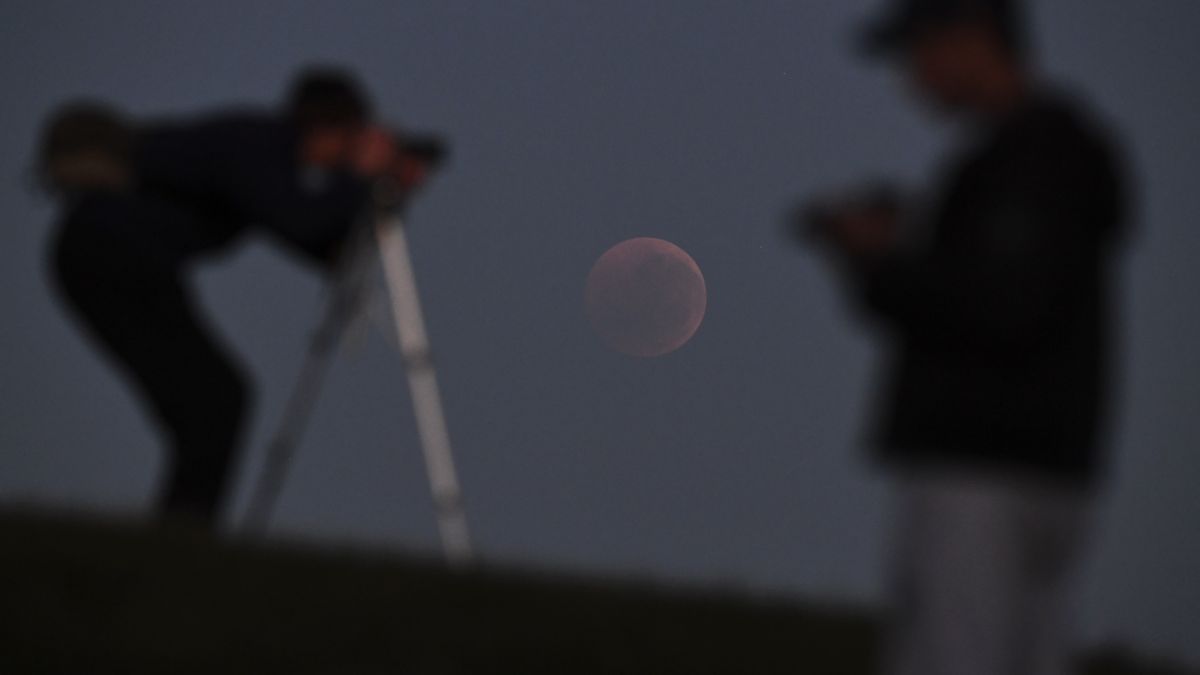 Osoba pochyla się, by spojrzeć przez obiektyw aparatu, skierowany na nisko położony księżyc krwi