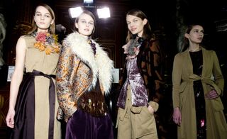 Three women, one wearing a nude dress with purple satin belt, one in a metallic orange coat with fur collar and one in khaki trousers, a padded purple velvet waistcoat and orange and black coat