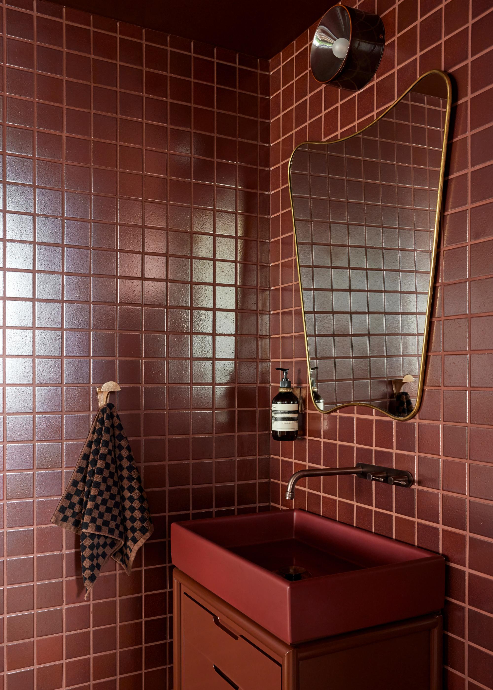 A small bathroom with maroon red tiles, a large mirror, a checkerboard hand towel, a red basin, and a set of drawers