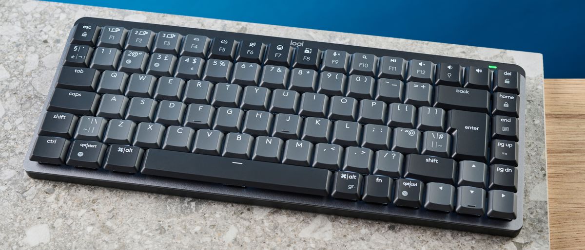 A photo of the Logitech MX Mechanical Mini keyboard in black and gray, on a stone slab and wooden table with a blue background.