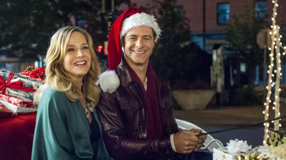 A man and woman sit in a sleigh filled with presents the man wears a santa hat