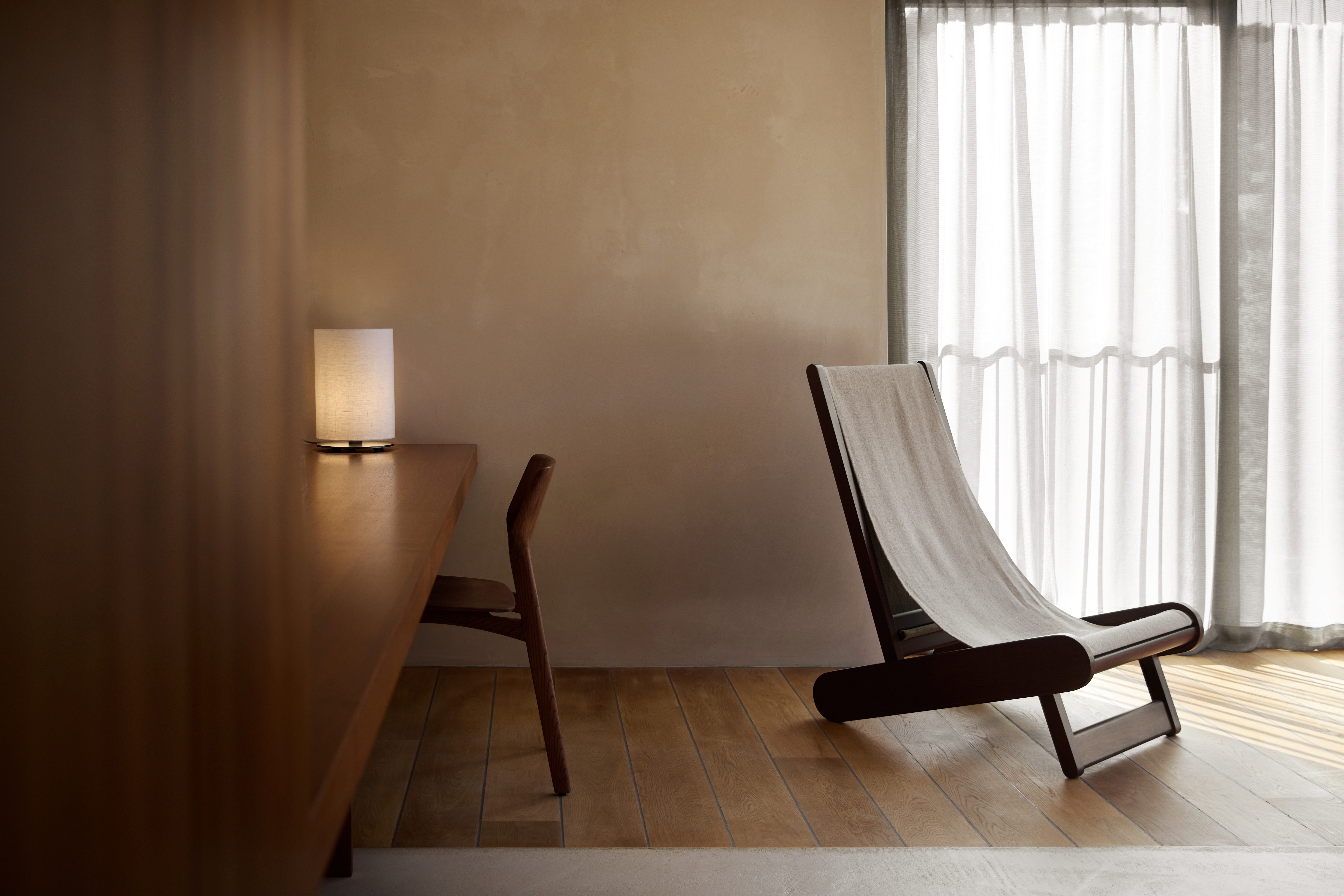 A minimalist hotel room with wooden floors and closets, white curtains, and wood furnishings mixed with neutral textiles.