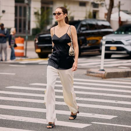 Woman wears black satin tank, white pants, and heeled sandals while walking on a cross walk.