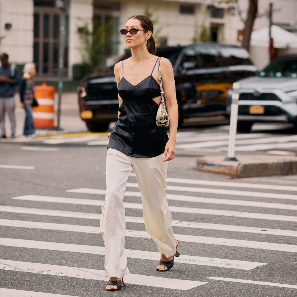 Woman wears black satin tank, white pants, and heeled sandals while walking on a cross walk.