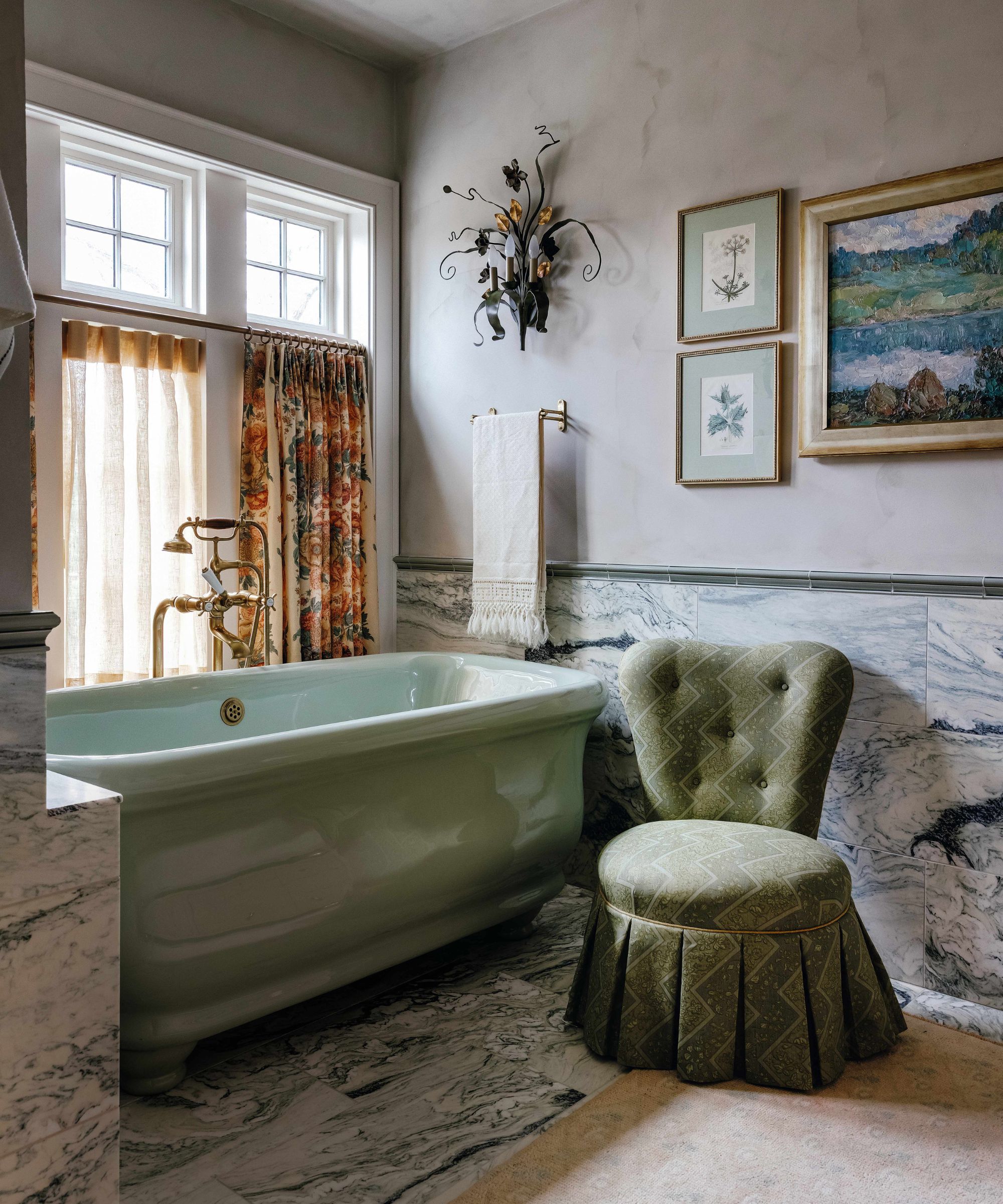 a layered bathroom with green marble half height walls and floor tiles, green bath tub and slipper chair with floral light, art, and cafe curtain