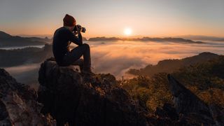 Person sitting with the back to the camera on a stone on a mountain, looking through the viewfinder of the camera, capturing the sunset 