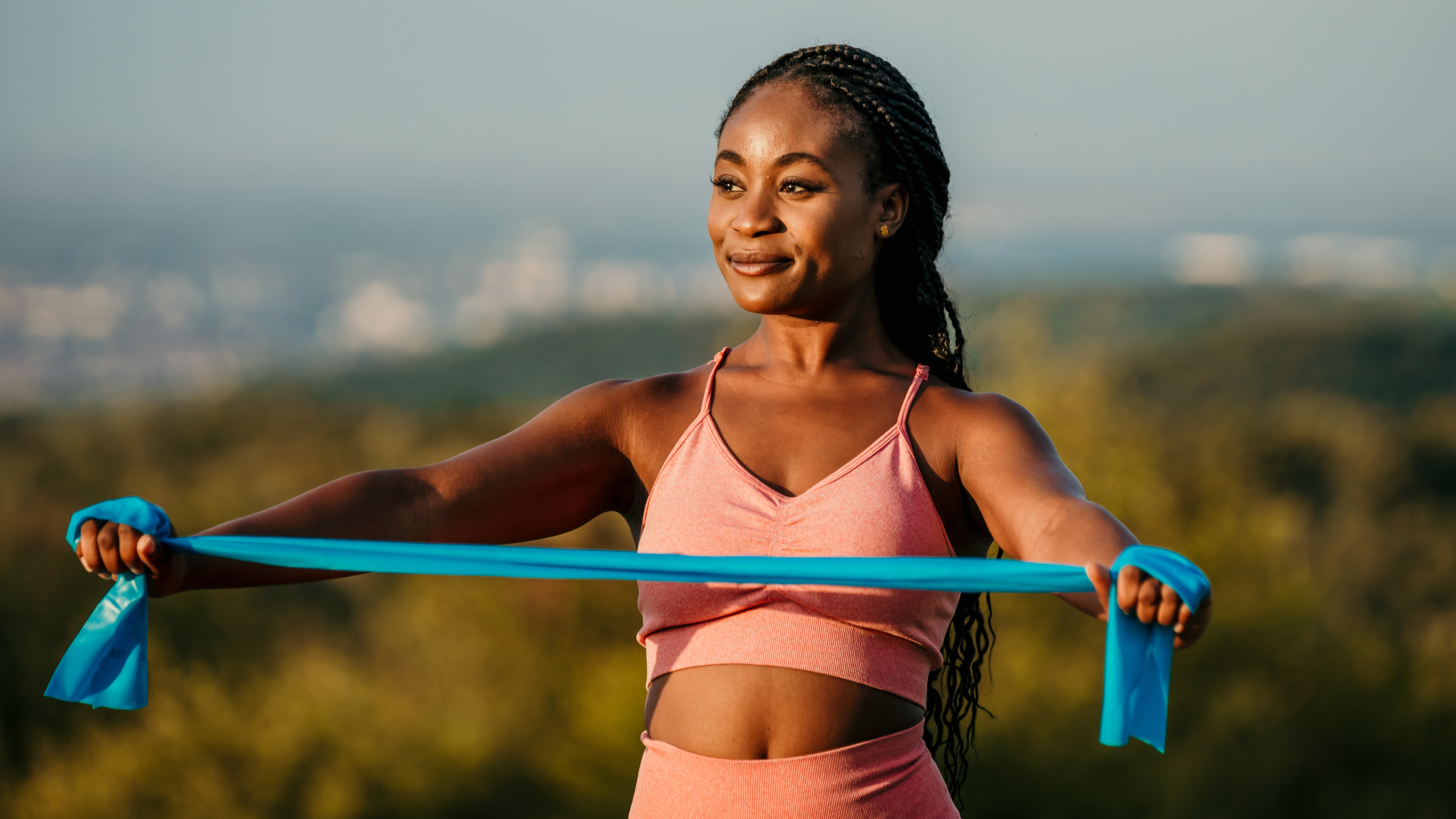 A woman stands holding a resistance band taut between her hands, with her arms long and elbows slightly bent. Behind her we see a blurred city skyline.