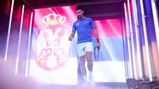 Novak Djokovic walks out onto a tennis court in front of a large screen depicting the flag of Serbia