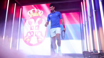 Novak Djokovic walks out onto a tennis court in front of a large screen depicting the flag of Serbia