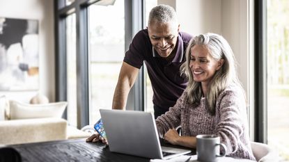 A smiling older couple look at a laptop together at their dining room table.