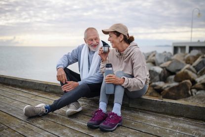 Active senior couple runners taking break during doing exercise outside on the pier by the sea, drinking tea