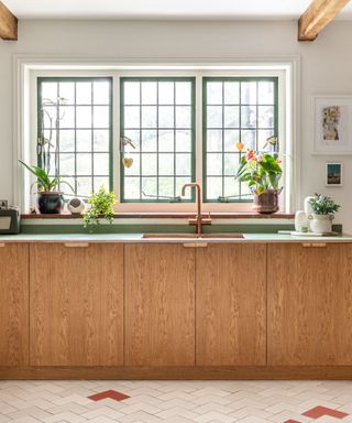 A kitchen with three green windows, plants on the windowsill, a green countertop and copper sink, light wooden cabinets and white tile floors.