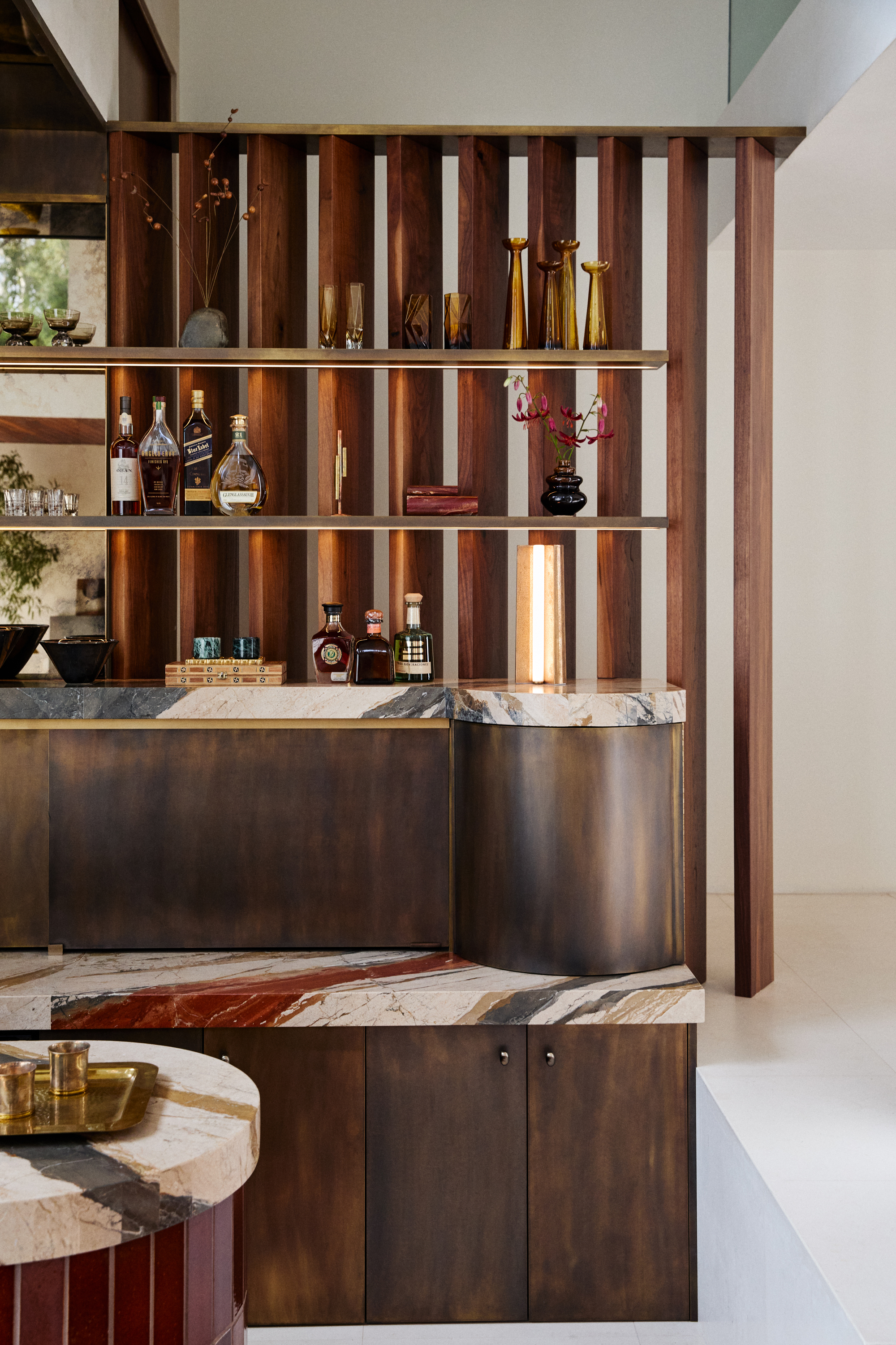 close up of sunken home bar with brass cabinetry, marble countertops, a finned walnut screen divider with built-in shelves styled with bottles and vases