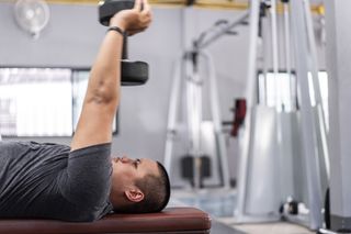 Man lying on weight bench holds dumbbell in two hands above his head