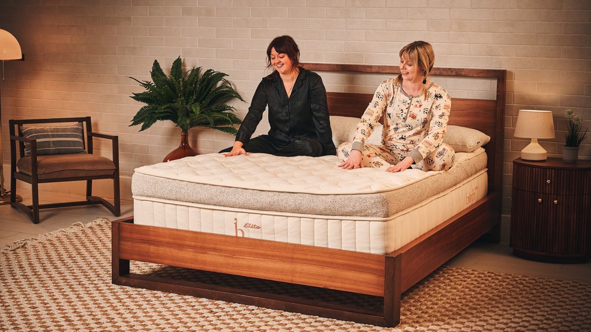 Two mattress testers sit on top of the beige and brown Birch Elite Natural mattress, which is placed on a wooden bed frame inside a neutrally decorated bedroom