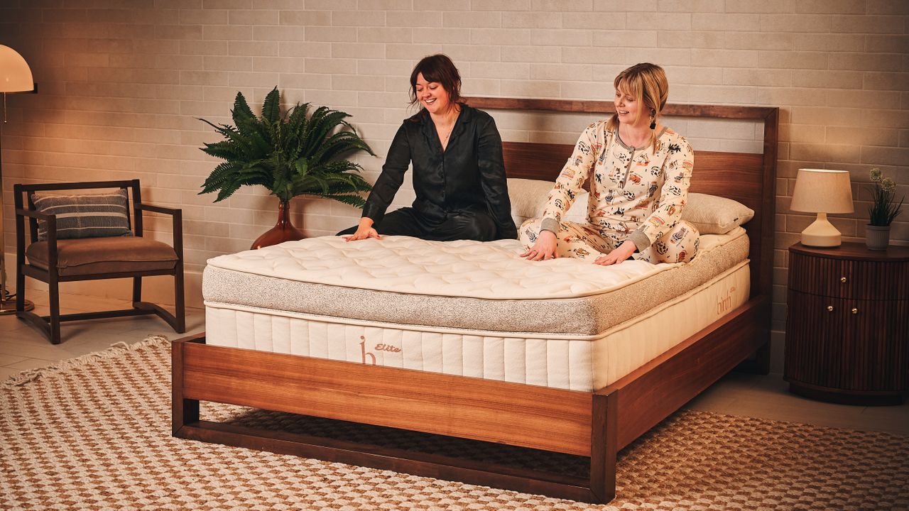 Two mattress testers sit on top of the beige and brown Birch Elite Natural mattress, which is placed on a wooden bed frame inside a neutrally decorated bedroom
