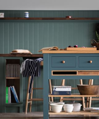 A dark green kitchen with a dark green kitchen island with a wooden surface, drawers, and wooden shelves with bowls and books on the lower