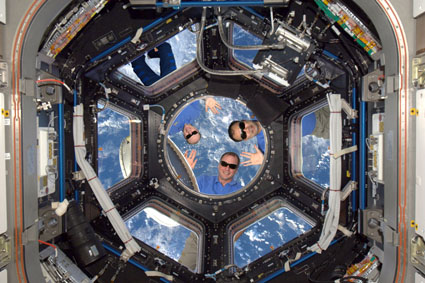 Les trois astronautes à l'extérieur de la Station spatiale internationale portent des lunettes de soleil.  Ils sourient et saluent la caméra.