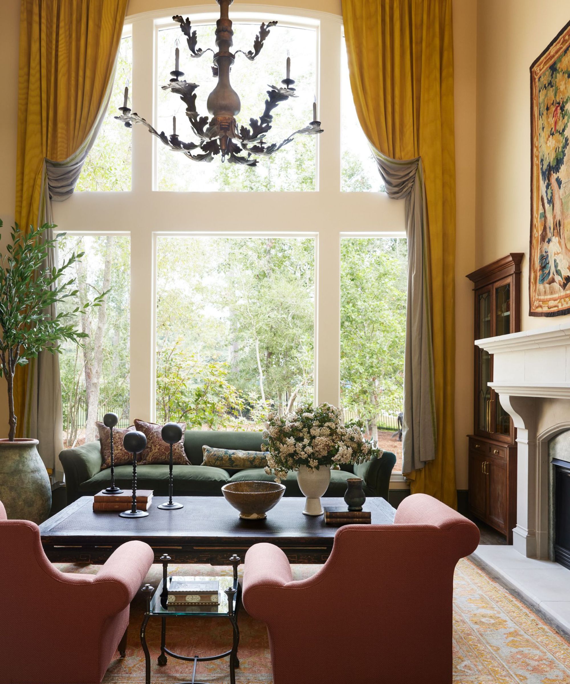 A traditional living room with a high ceiling, neutral walls, and large floor-to-ceiling windows with dark ochre curtains. An ochre-patterned rug, two pink armchairs, a black coffee table, and a dark green sofa.