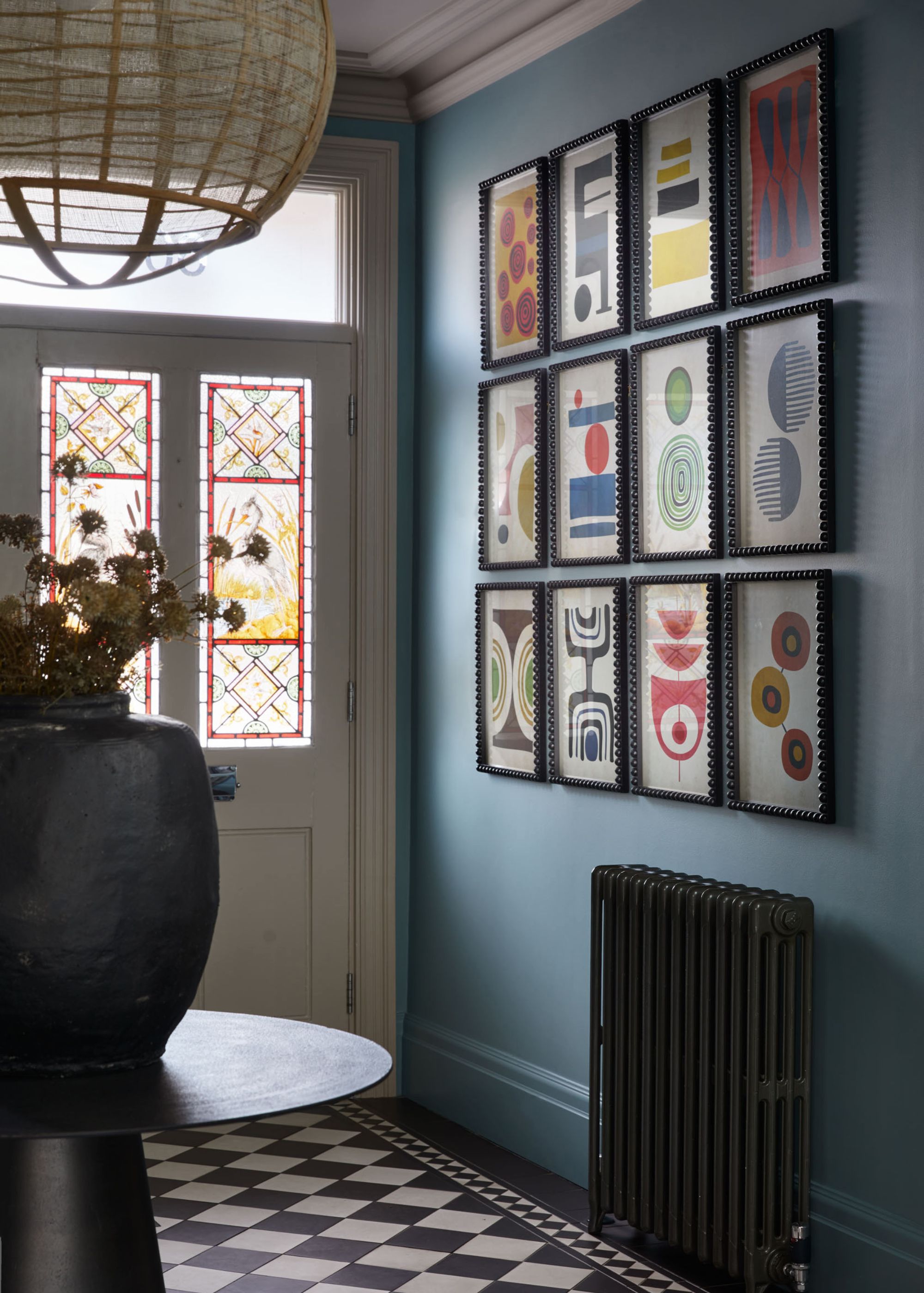 An entryway in a traditional home with blue walls, a white front door with stained glass, a gallery wall of abstract, colorful art, a black column radiator, and black and white tiled flooring.