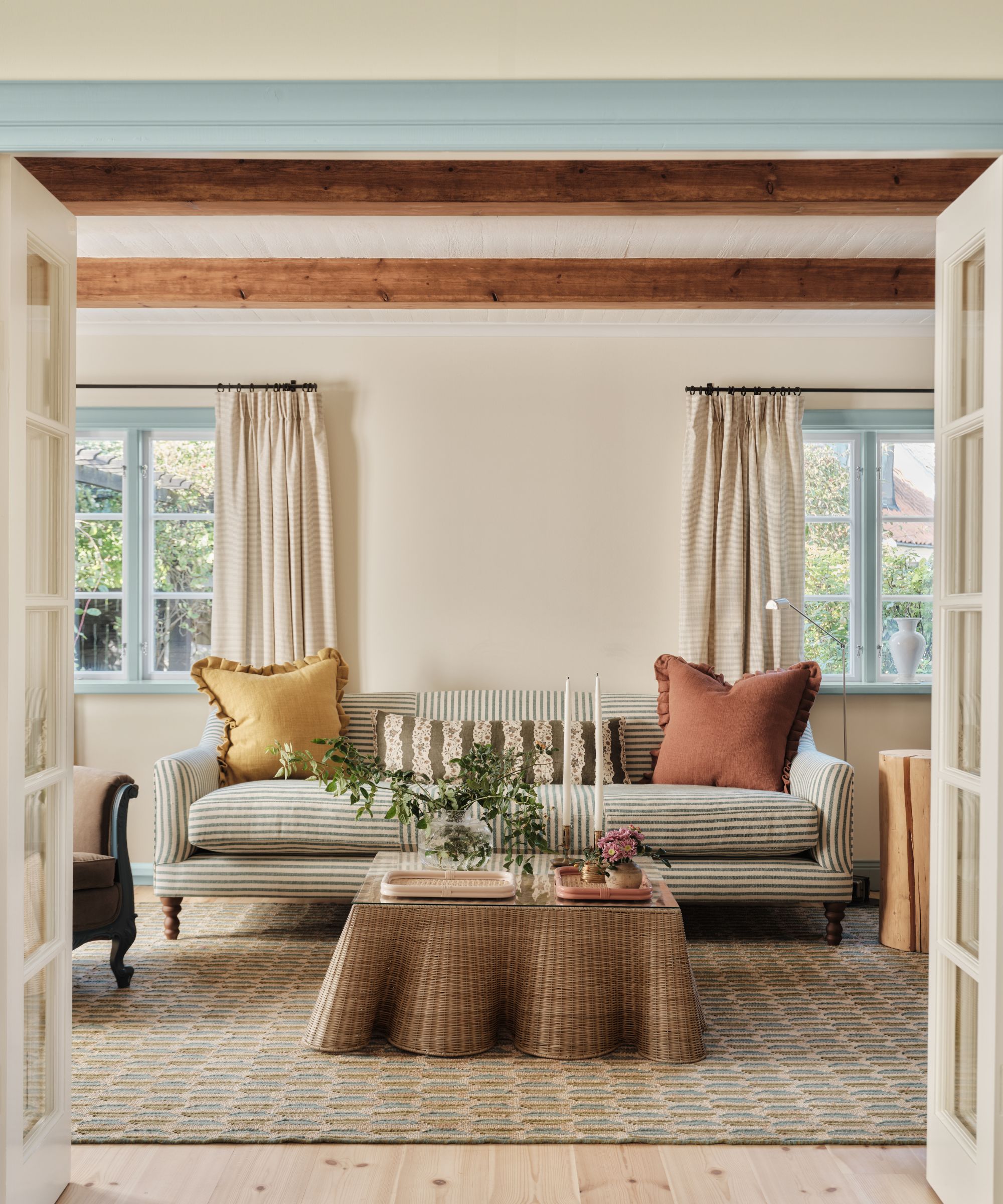 Pale cream living room with wooden beams and window frames painted in pale blue. A striped sofa sits in the middle with a rattan coffee table in front