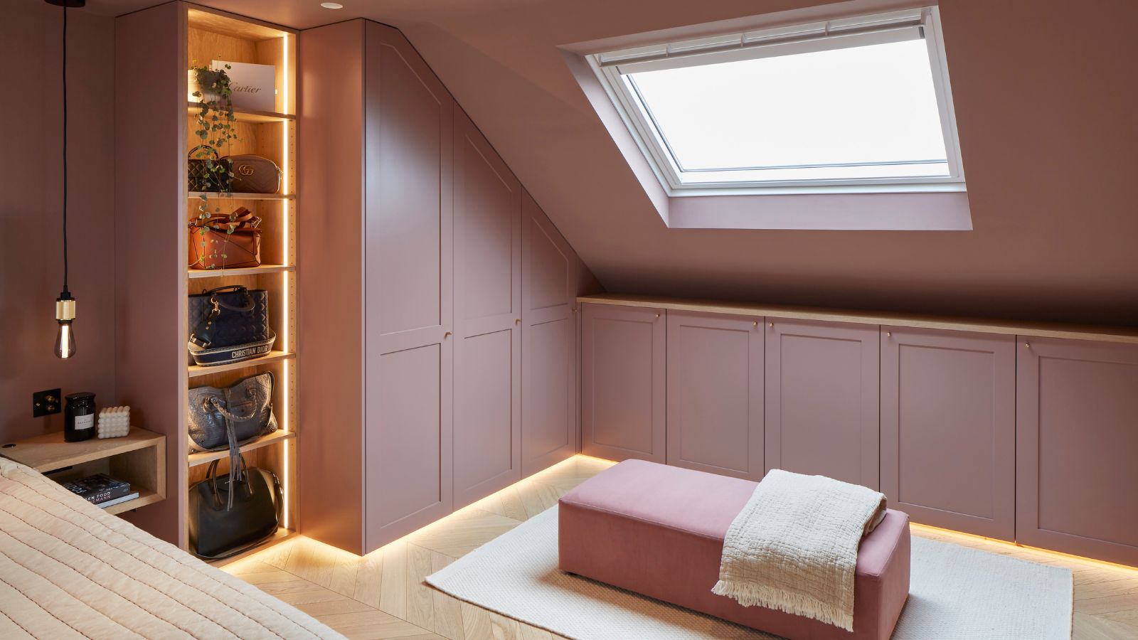 A softly lit loft dressing room with sloped ceilings and a large rectangular skylight bringing in natural light. The cabinetry, walls and built-in wardrobes are painted in a muted dusty pink tone, creating a cohesive, cocooning feel. Along one wall, low fitted cupboards run beneath the slanted ceiling, with subtle under-cabinet lighting casting a warm glow along the floor.
