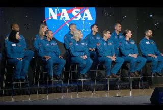 11 people in blue flight suits sit on a stage with a projection of nasa's logo on the wall behind them