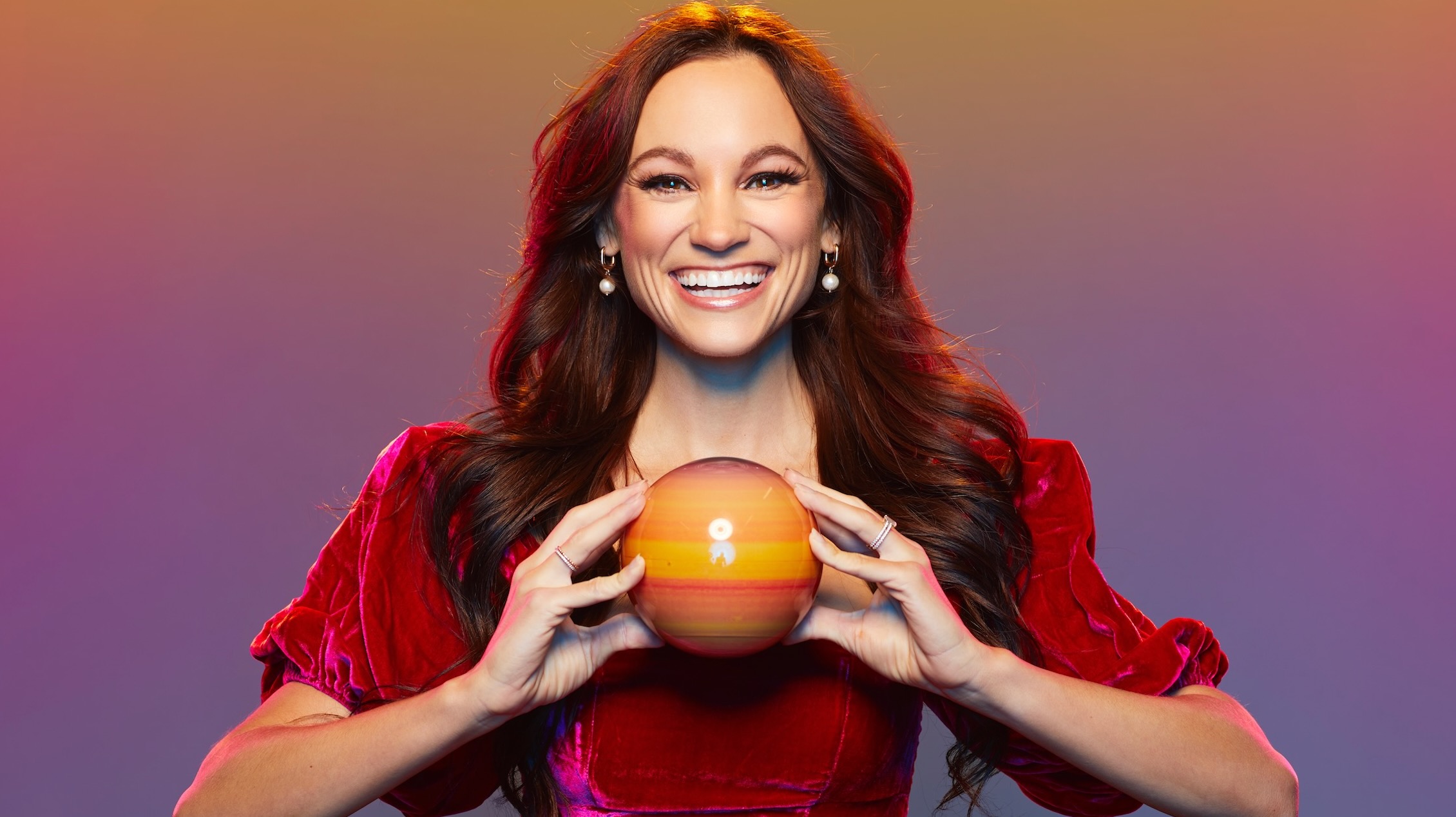 a smiling brown-haired woman in a red dress holds a shiny orange-red sphere that's about the size of a grapefruit