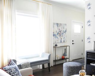 White lounge with custom made floor length drapes, window bench, gray pouffe, and entrance console with wall art.