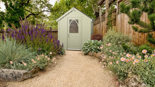 Sage green shed in a garden with plants on either side of a gravel walkway