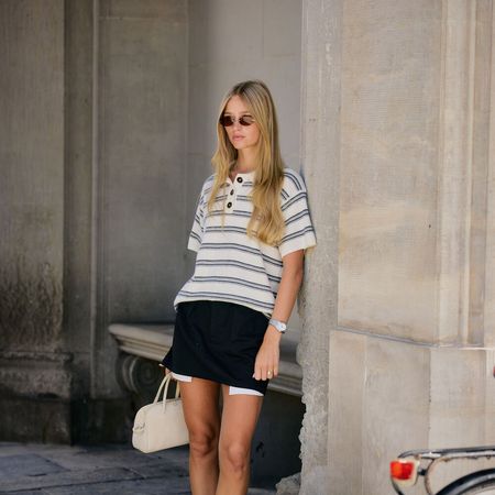 Woman wears striped top, black mini skirt, pointed toe heels, and a white handbag while leaning on a wall. 