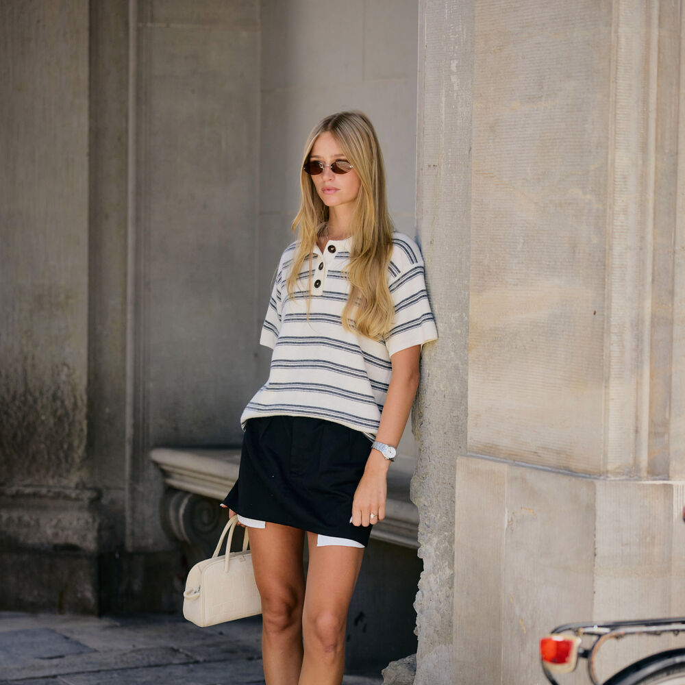 Woman wears striped top, black mini skirt, pointed toe heels, and a white handbag while leaning on a wall. 