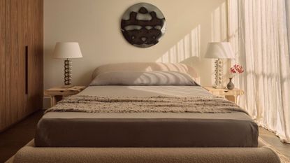 A white bedroom with a large bed in gray bedding, a textured coverlet, wood nightstands, matching textured lamps, a vase, and a round hammered chrome wall hanging
