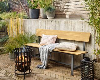A wooden bench with outdoor cushion, throw and black metal firepit