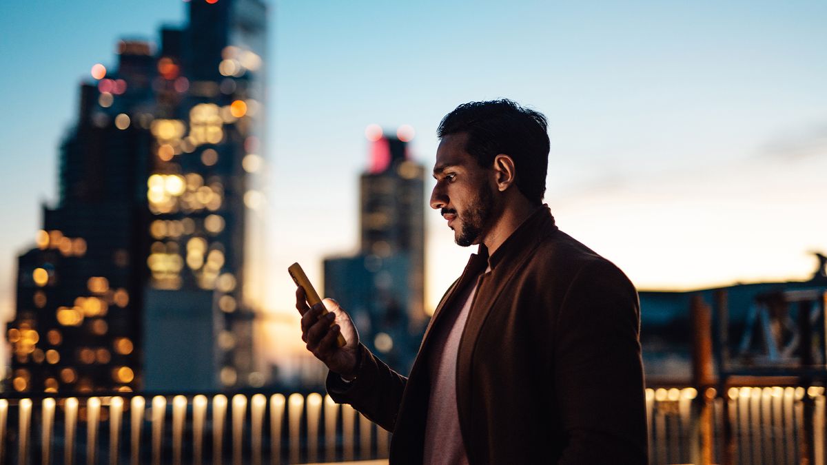 Tech workers concept image showing a man looking at job opportunities on a smartphone on a rooftop at sunset with buildings in background.